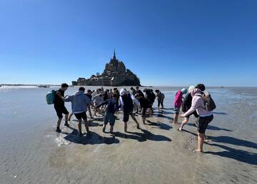 Das Bild zeigt eine Gruppe von Jugendlichen, die auf dem Meeresgrund vor dem Mont-Saint-Michel spazieren gehen. Der Strand ist bei Ebbe trocken gefallen und die Gruppe ist auf dem Weg zum Felsen. Im Hintergrund ist der Mont-Saint-Michel deutlich sichtbar, unter einem strahlend blauen Himmel. Die Stimmung ist fröhlich und abenteuerlich