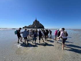 Das Bild zeigt eine Gruppe von Jugendlichen, die auf dem Meeresgrund vor dem Mont-Saint-Michel spazieren gehen. Der Strand ist bei Ebbe trocken gefallen und die Gruppe ist auf dem Weg zum Felsen. Im Hintergrund ist der Mont-Saint-Michel deutlich sichtbar, unter einem strahlend blauen Himmel. Die Stimmung ist fröhlich und abenteuerlich