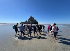 Das Bild zeigt eine Gruppe von Jugendlichen, die auf dem Meeresgrund vor dem Mont-Saint-Michel spazieren gehen. Der Strand ist bei Ebbe trocken gefallen und die Gruppe ist auf dem Weg zum Felsen. Im Hintergrund ist der Mont-Saint-Michel deutlich sichtbar, unter einem strahlend blauen Himmel. Die Stimmung ist fröhlich und abenteuerlich